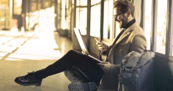 man sitting near window holding phone and laptop