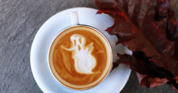 white ceramic teacup filled with ghost illustration coffee latte on white ceramic saucer beside maroon leaf photography