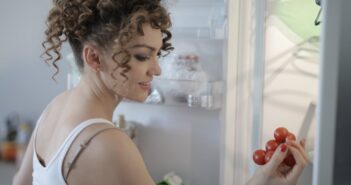 Side view of content female in casual wear standing in kitchen near opened fridge and picking fruit for breakfast