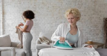 Busy woman ironing clothes and black girlfriend folding clothes during daily routine at home