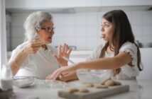 Calm senior woman and teenage girl in casual clothes looking at each other and talking while eating cookies and cooking pastry in contemporary kitchen at home