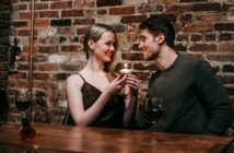 Side view of loving couple sitting near brick wall at wooden table with cupcake with burning candle looking at each other while celebrating anniversary in cafe