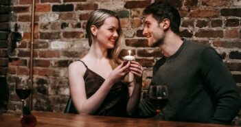 Side view of loving couple sitting near brick wall at wooden table with cupcake with burning candle looking at each other while celebrating anniversary in cafe