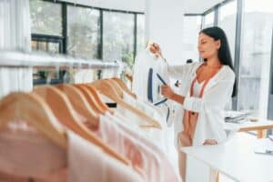 Irons clothes. Seamstress is in her office that is with different clothes