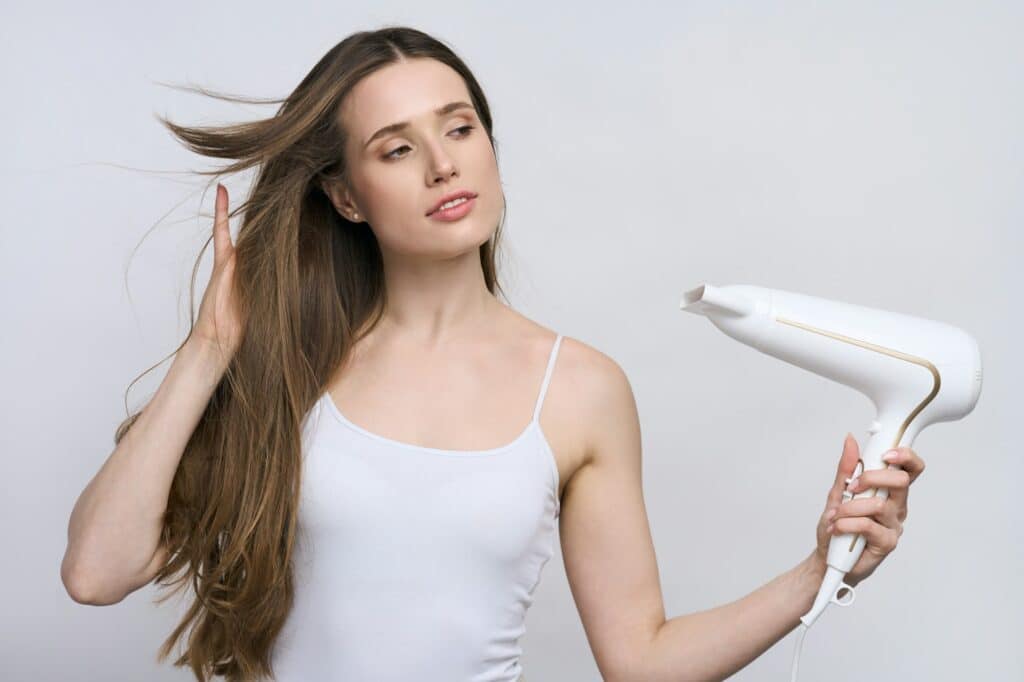 Isolated portrait on white backdrop of young woman blowing warm air fron hair dryer on long wet hair