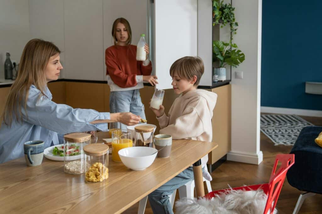 Loving teenage daughter holding bottle of milk from fridge for family breakfast with mom and brother