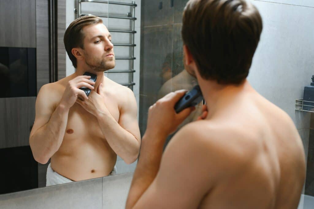 Reflection of young man in mirror shaving with electric shaver