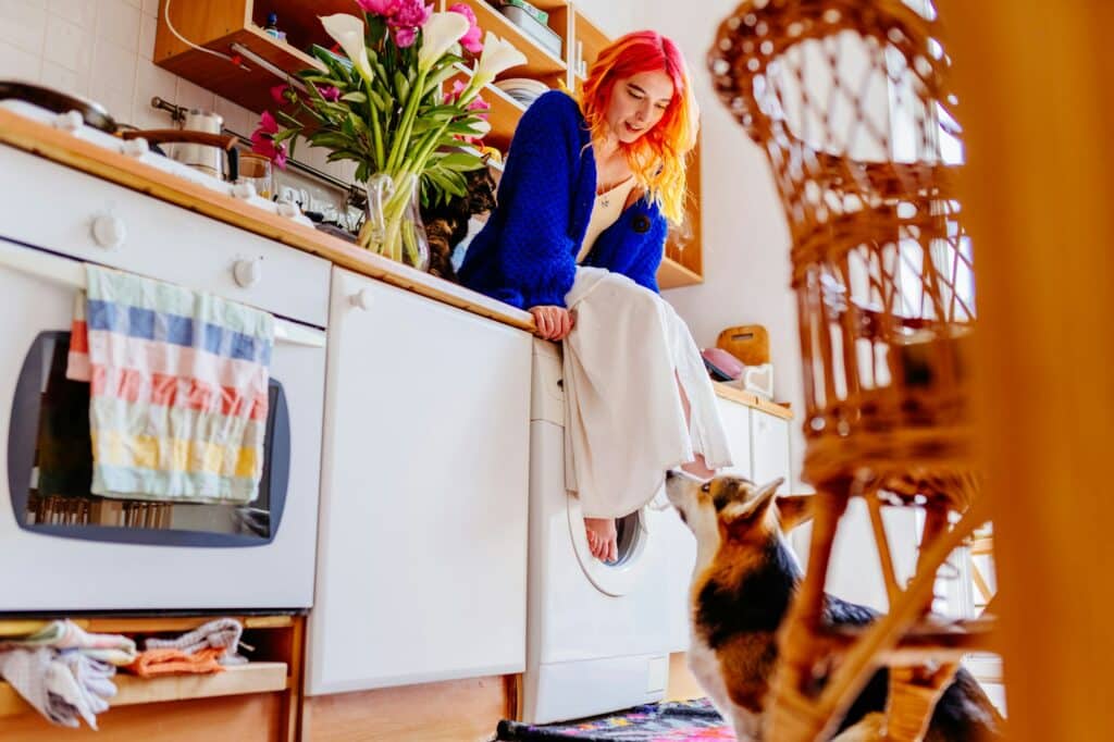 Caucasian woman with vibrant red hair sitting on washing machine in cozy kitchen.