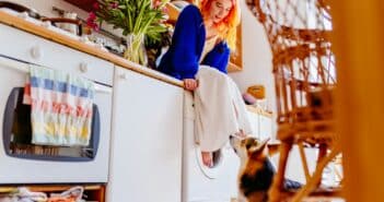 Caucasian woman with vibrant red hair sitting on washing machine in cozy kitchen.