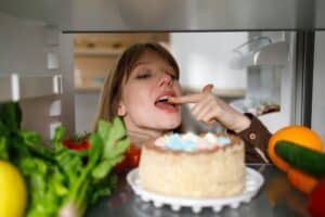 Cheerful woman tasting cake with finger in fridge