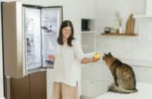 Cute cat smelling apple in woman hand on background of opened fridge