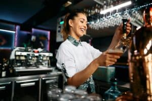 Happy female barista pouring beer from a beer tap while working in pub.