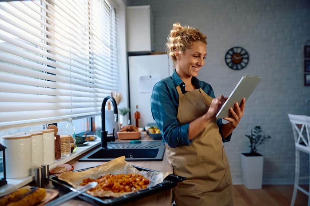 Happy woman reading a recipe on touchpad while preparing food in the kitchen.