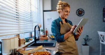 Happy woman reading a recipe on touchpad while preparing food in the kitchen.