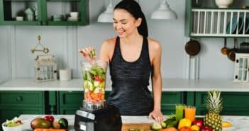 Healthy slim girl adding cucumber to the blender full of fresh vegetables for making healthy