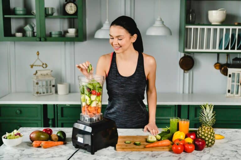 Healthy slim girl adding cucumber to the blender full of fresh vegetables for making healthy