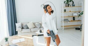 In headphones and with vacuum cleaner. Woman in casual clothes is indoors at home