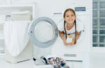 Smiling female child with glad expression, poses in washing machine, holds detergent