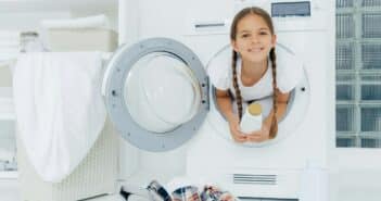 Smiling female child with glad expression, poses in washing machine, holds detergent