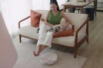 Woman relaxing on couch with dog and robotic vacuum cleaner.