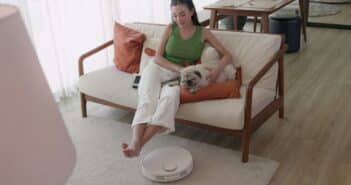 Woman relaxing on couch with dog and robotic vacuum cleaner.