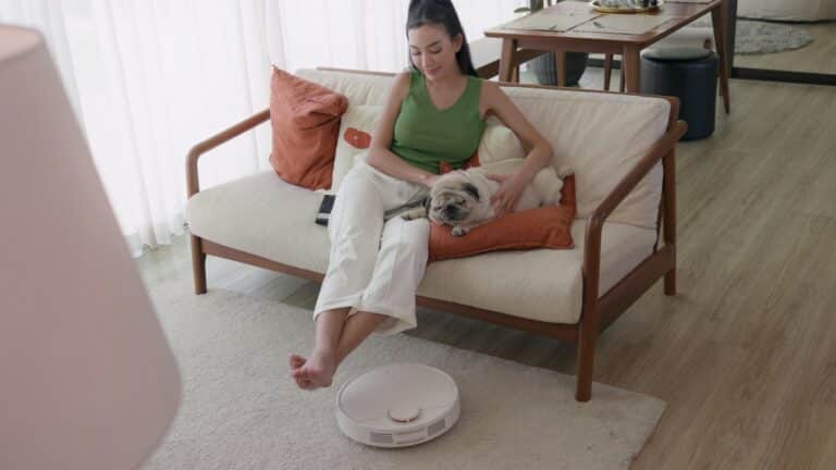 Woman relaxing on couch with dog and robotic vacuum cleaner.