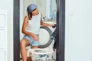 Woman sits in front of washing machine and loads it with dirty laundry.