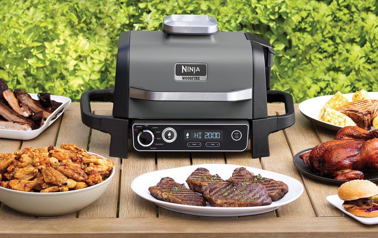 Un barbecue électrique de la marque Ninja Kitchen posé sur une table de jardin à côté d'assiettes de viandes grillées.