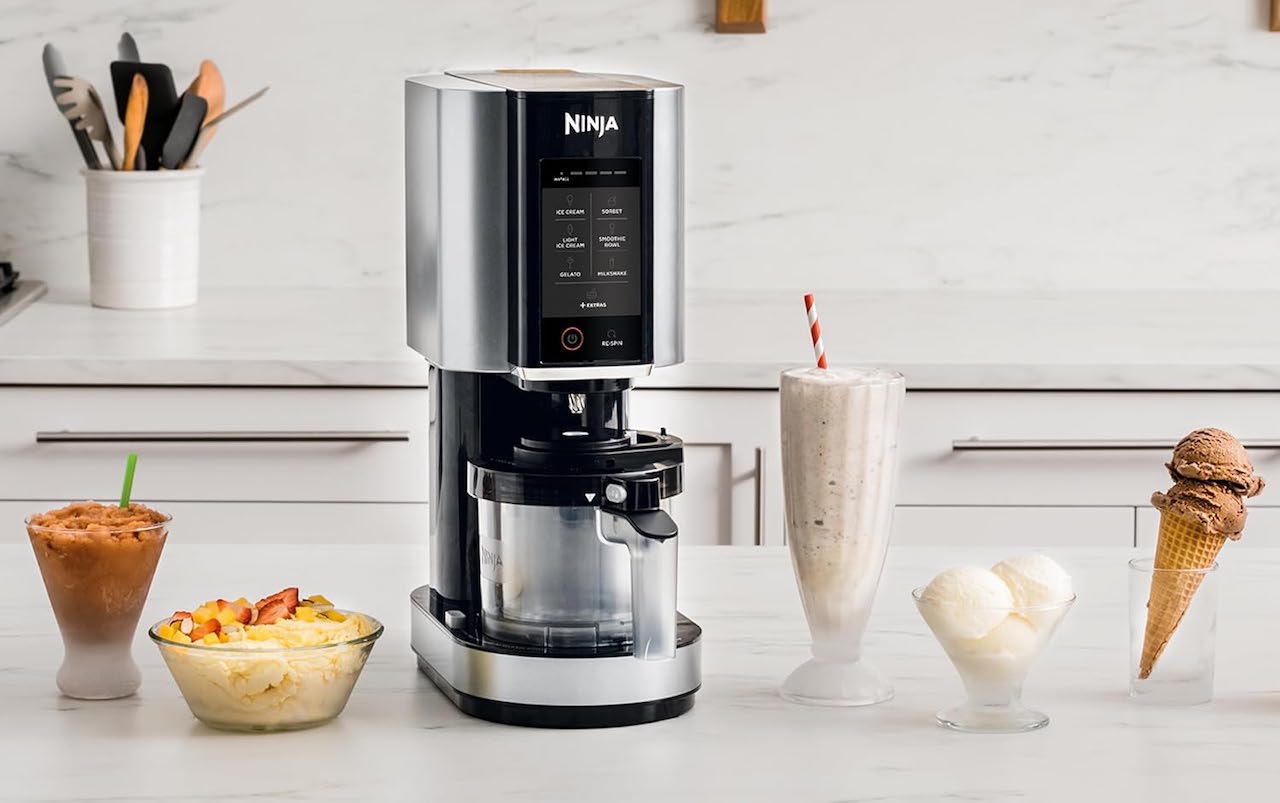 Une machine à desserts glacés de la marque Ninja Kitchen. La machine est posée sur un plan de travail et à côté sont disposés des desserts glacés : milkshakes, glaces, etc..