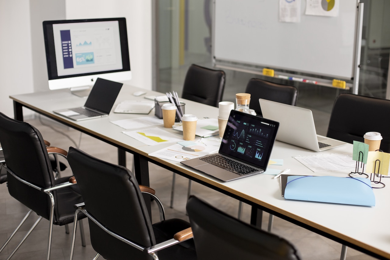 Une salle de réunion pour le travail. Sur la table on voit des ordinateurs, des tasses, des pochettes et feuilles.